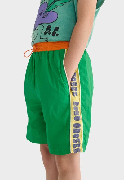 Enfant portant un short vert avec une ceinture orange et une bande latérale jaune portant le texte violet "BOBO CHOSES", la main dans la poche.
