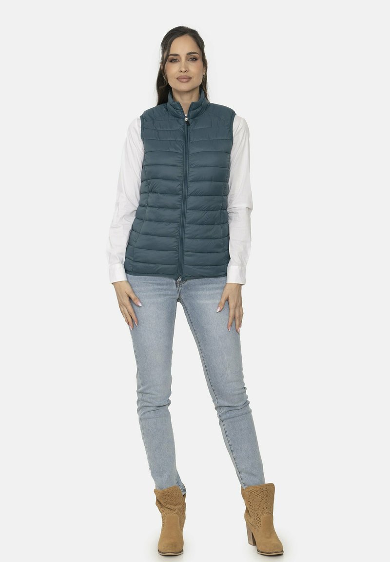 Chaleco acolchado de color verde azulado oscuro con textura de acolchado vertical, cremallera frontal y cuello alto, combinado con una camiseta blanca y jeans de color azul claro.