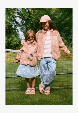 Kaks last kannavad sobitatud rõivaid, mis sisaldavad pehmet roosat fliisjopet südamemustriga, helesiniseid teksaseid südamekujundusega ja sandaale.