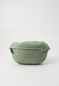 Grüne Stofftasche mit runder Form, ausgestattet mit einem Logo-Patch auf der Vorderseite und einem Reißverschluss. Texturiertes Gewebe und genähte Details.