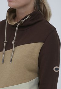 Hoodie color block marrone e beige con colletto alto, cordini intrecciati e patch con logo. Tessuto morbido con vestibilità rilassata.