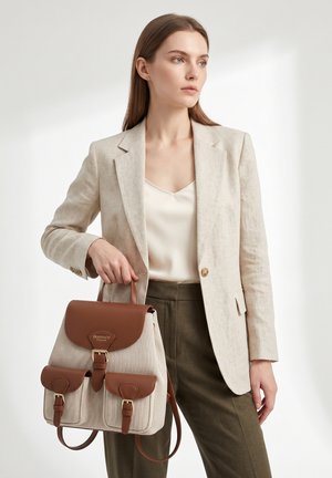 Vrouw in beige blazer en wit topje houdt een beige en bruine rugzak met leren accenten en voorzakken vast, staand tegen een lichte achtergrond.