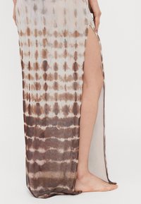 Long, sheer skirt with a tie-dye pattern in brown and beige. Features a high side slit, smooth texture, and a flowy design.