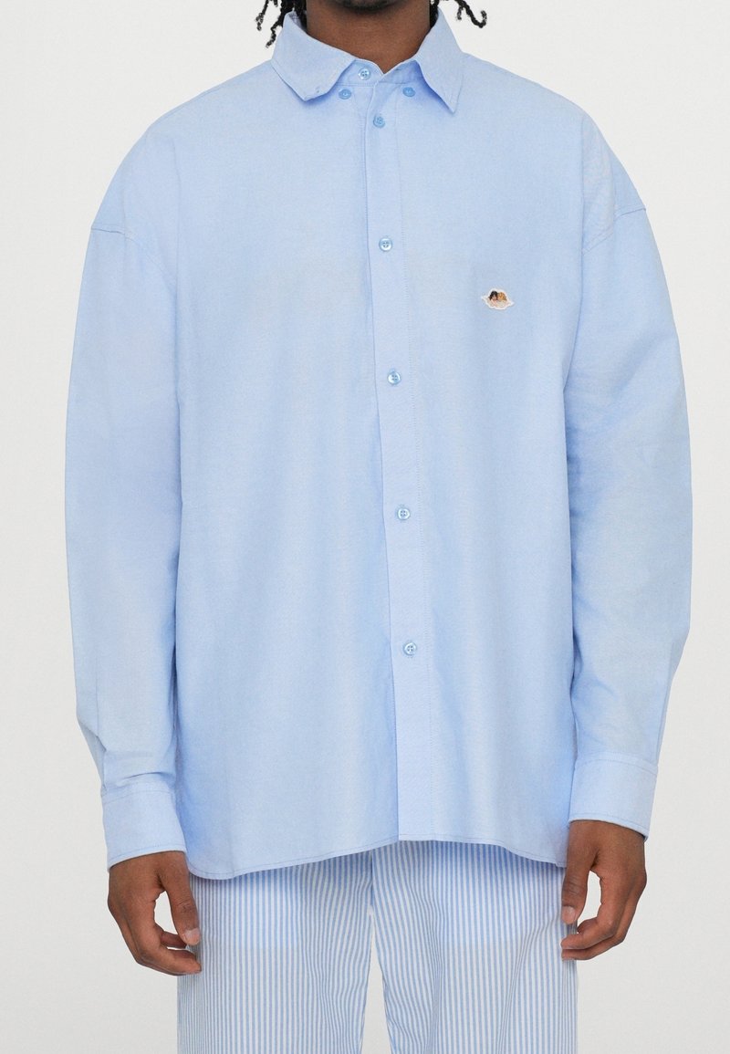 Man wearing loose light blue button-up shirt with small embroidered patch on chest, paired with light blue and white striped pants.