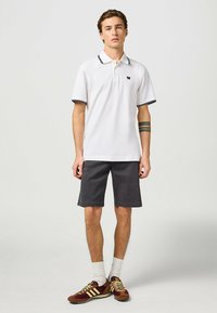 Polo bianco con rifiniture nere, abbinato a pantaloni corti grigi scuri. Il modello indossa sneakers marroni e gialle con calze bianche. Design semplice, vestibilità casual.