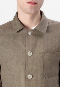 Brown linen button-up shirt, featuring a classic collar, three buttons, and a chest pocket. Textured fabric with a subtle pattern.