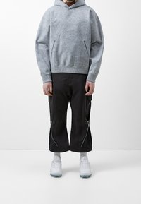 Grå stickad hoodie med en strukturerad yta, har en känguruficka. Matchas med svarta korta byxor med dragkedja och vita träningsskor.