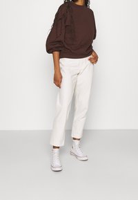 Hnědá mikina s nabíranými rukávy, bílé kalhoty s vysokým pasem a bílé vysoké tenisky. Jednoduchý, pohodlný outfit v neutrálních tónech.