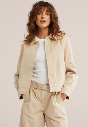 Ženska z kodrastimi lasmi, oblečena v bež srajčno jakno s gumbi, belo rebrasto majico in bež hlače z visokim pasom ter pasom, z rokami v žepih.