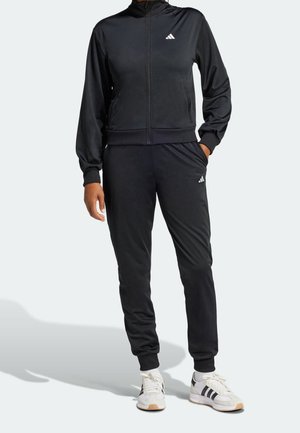 Persoon die een zwart Adidas trainingspak draagt met een jas met rits en een broek, gecombineerd met witte Adidas sneakers met zwarte strepen.