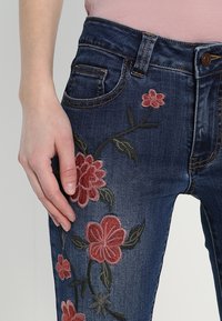 Tmavě modré džínové kalhoty s květinovou výšivkou v červené a růžové barvě. Mají klasický pětikapsový design a zapínání na zip s knoflíkovým uzávěrem.