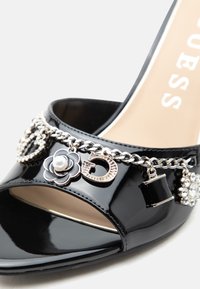 Chaussure noire en cuir verni à bout ouvert avec une chaîne argentée comportant une fleur, la lettre D et des charms sac à main sur la bride avant.