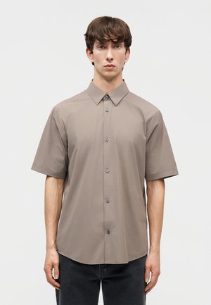 Jeune homme aux cheveux bruns courts portant une chemise taupe à manches courtes et un pantalon foncé, sur un fond uni.