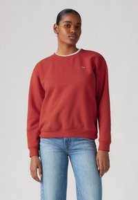 Sweat-shirt à col rond rouge en tissu doux, avec une bordure de col blanche et un petit logo sur la poitrine. Assorti avec un jean bleu.
