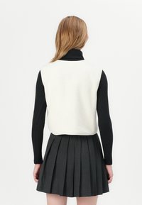 Weißer Fleece-Weste mit einem ärmellosen Design, kombiniert mit einem schwarzen Langarmoberteil. Ein grauer Faltenrock vervollständigt das Outfit.