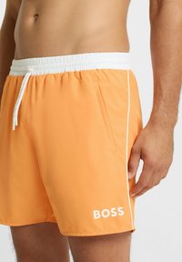 Oranje zwembroek gemaakt van lichtgewicht stof met een elastische witte tailleband, trekkoord en zijzakken. Voorzien van een wit logo en witte biesdetail.