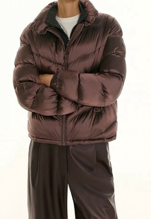 Personne portant une doudoune marron foncé brillante, les bras croisés sur une chemise blanche et un pantalon large marron foncé.