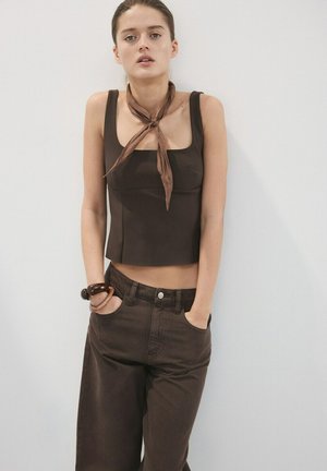 Mladá žena oblečená v hnedom bezrukávovom topu, zodpovedajúcich širokých nohaviciach, šatke na krku a náramkoch stojí s rukami v vreckách oproti jednému jednoduchému múru.