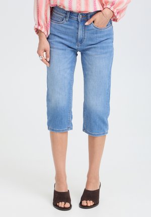 Donna che indossa jeans in denim azzurro chiaro corti, camicetta rosa a righe, sandali mule marroni aperti in punta con tacco e anelli d'argento sulla mano sinistra.