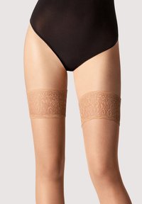 Beige lynlår strømper med blonder og glat tekstur samt blomstermønster øverst, båret over et sort shapewear beklædningsstykke.