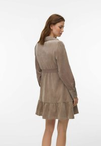 Femme portant une robe taupe à manches longues avec un ourlet froncé et une taille smockée, en pose de dos aux trois quarts sur un fond uni.