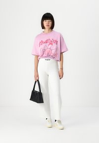 Pink oversized T-shirt with graphic design, white high-waisted trousers, and black handbag. White sneakers with a yellow sole in the foreground.