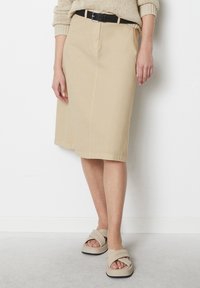 Beige knielange Rock mit Vorderfalten, aus glattem Stoff gefertigt, kombiniert mit einem schwarzen Gürtel. Getragen mit beigen Sandalen mit breiten Riemen.