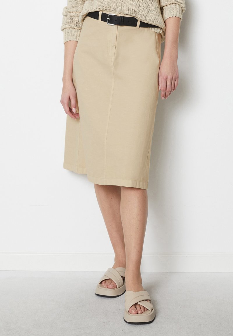 Beige knielange Rock mit Vorderfalten, aus glattem Stoff gefertigt, kombiniert mit einem schwarzen Gürtel. Getragen mit beigen Sandalen mit breiten Riemen.