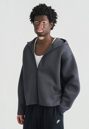 Szara bluza z kapturem na suwak, miękka w dotyku, duże przednie kieszenie oraz luźny krój, zestawiona z czarnymi spodniami dresowymi z logo.