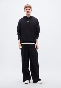 Μαύρο φούτερ με κουκούλα, μπροστινή τσέπη και κορδόνι, συνδυασμένο με άνετο μαύρο παντελόνι. Παπούτσια είναι λευκά αθλητικά. Απλός, μινιμαλιστικός σχεδιασμός.