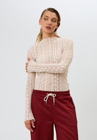 Tynn langermet topp i lys beige med blonde og stripemønster; kombinert med avslappede maroonbukser med elastisk liv og snor.