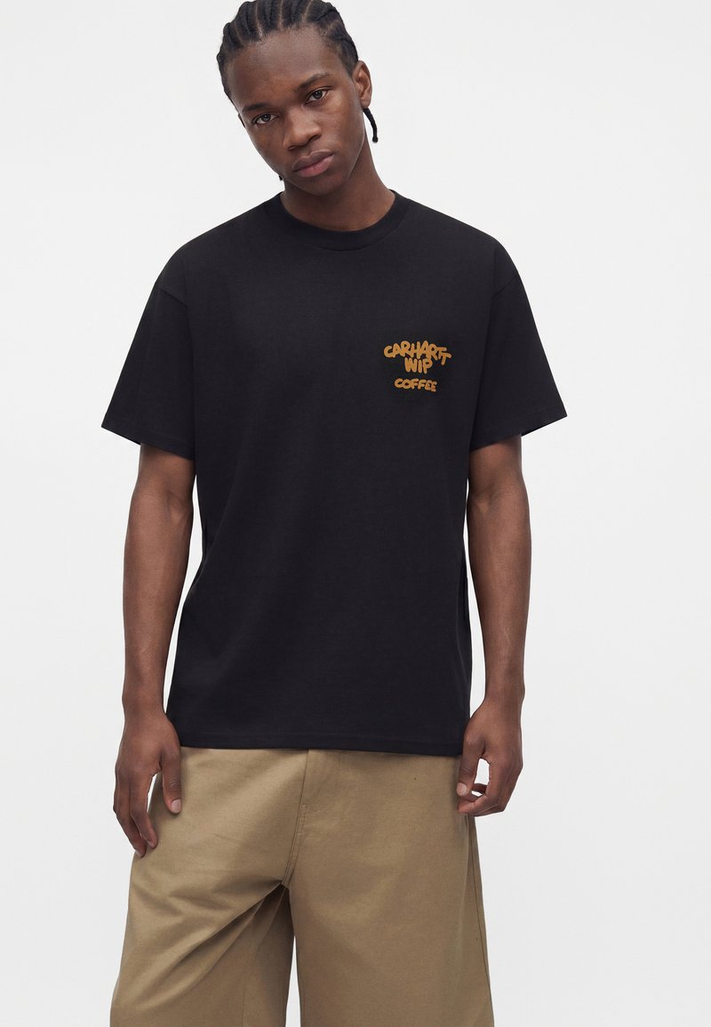 Mann mit geflochtenen Haaren, der ein schwarzes Carhartt WIP Coffee T-Shirt und beigefarbene Hose trägt, vor einem schlichten weißen Hintergrund stehend.