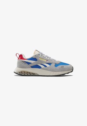 Zapatilla Reebok con tela azul, superposiciones de ante gris, entresuela blanca con diseño de panal, lengüeta trasera roja y logotipo de Reebok en blanco en el lateral.