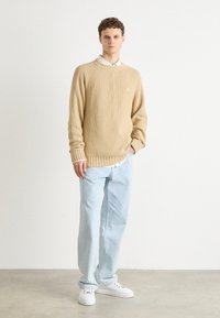 Beige ribbed sweater with a round neckline, worn over a white shirt, paired with light blue jeans and white sneakers. Simple design, relaxed fit.