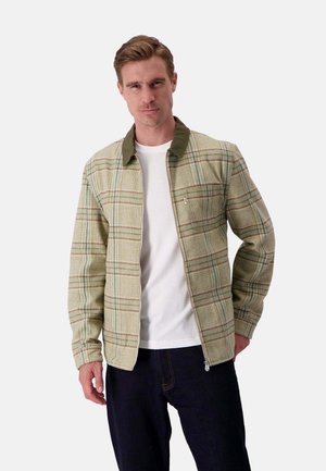 Grüne karierte Shirtjacke mit Kragen, mit beigen, braunen und hellblauen Streifen. Getragen über einem weißen T-Shirt, mit dunkler Jeanshose.