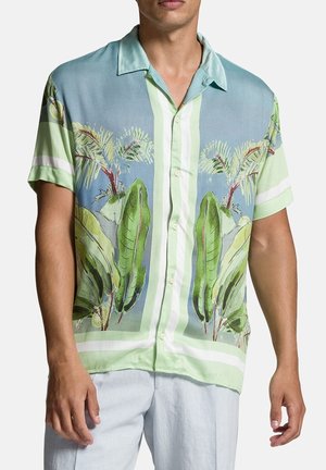 Mężczyzna w koszuli na guziki z krótkim rękawem, ozdobionej zielono-niebieskim tropikalnym wzorem liści i palm, w połączeniu z jasnymi spodniami.