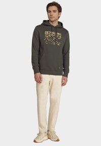 Szary hoodie z żółtym napisem, kangurzą kieszenią i ściągaczami, w połączeniu z kremowymi spodniami i beżowymi trampkami.