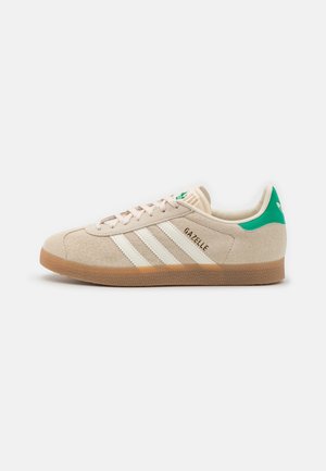 Beige Adidas Gazelle sneaker met witte strepen, rubberen zool en groene hieltab, zijaanzicht op een witte achtergrond.