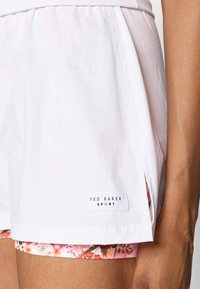 Baltas sporta šortus ar sānu šķirbiņu un ziedu apdruku iekšpusē. Zīmola etikete "TED BAKER SPORT" ir redzama apakšējā malā.