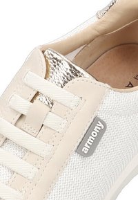 Zapatilla blanca con parte superior de malla texturizada, detalles de cuero beige, cordones planos, acabado metálico en la lengüeta y logotipo gris en el costado.