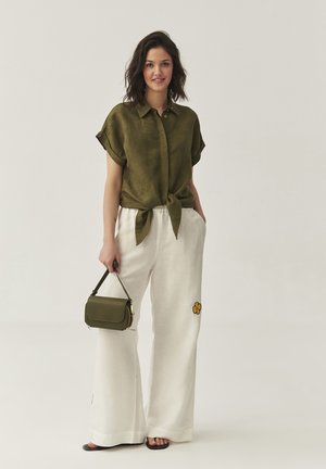 Chemise vert olive avec un nœud à l'avant, pantalon blanc à jambes larges avec broderie de fleurs jaunes, et un petit sac à main olive.