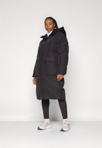 Schwarze Pufferjacke mit Reißverschluss, großen vorderen Taschen und Kapuze. Das Modell trägt schwarze Leggings und weiße Turnschuhe.