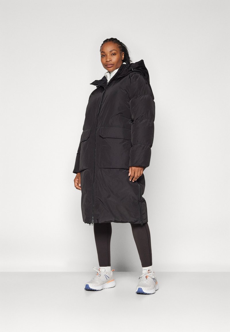 Schwarze Pufferjacke mit Reißverschluss, großen vorderen Taschen und Kapuze. Das Modell trägt schwarze Leggings und weiße Turnschuhe.