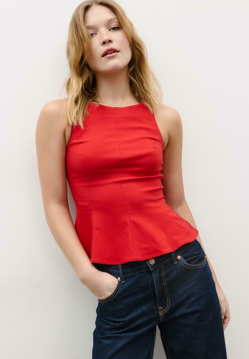 Mlada žena u crvenoj majici bez rukava s peplum opcijom i tamnoplavim trapericama, pozirajući s jednom rukom u džepu na jednostavnoj bijeloj pozadini.