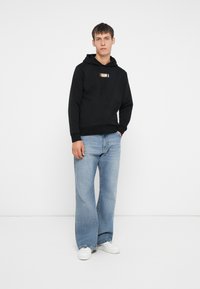 Felpa nera con cappuccio e cordini, con un logo frontale, abbinata a jeans a gamba larga blu chiaro e scarpe da ginnastica bianche, in piedi contro uno sfondo semplice.