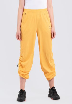 Pantalons de sport jaunes avec une surface texturée, taille élastique, poches latérales et détails de ourlet avec cordon de serrage. Portés avec des baskets noires.