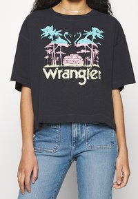 Sort cropped t-shirt med et farverigt grafisk motiv af palmetræer og flamingoer, med ordet "Wrangler" i lys gul nederst.