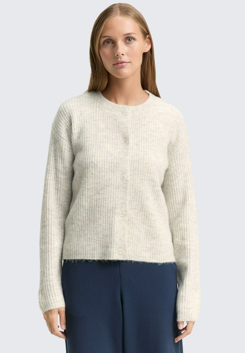 Femeie cu păr blond drept, purtând un cardigan crem cu nervuri și nasturi, și pantaloni bleumarin, stând în fața unui fundal deschis simplu.