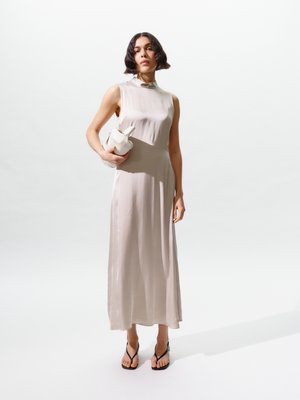 Vrouw in een mouwloze lichtbeige satijnen jurk die een witte opgevouwen clutch vasthoudt, draagt zwarte teenslippers en staat tegen een effen witte achtergrond.