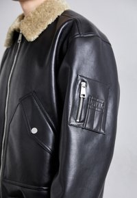 Fekete bőrkabát, műszőrme gallérral, cipzáras zsebbel az ujjon és ferde elülső zsebekkel, sima felülettel.